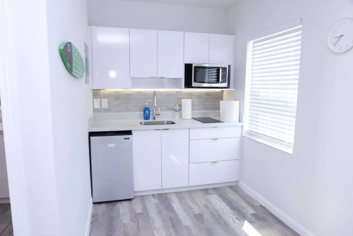 The image showcases a modern, compact kitchen area within a bright Miami studio apartment. The focal points include sleek white cabinetry, a stainless steel mini fridge, under-cabinet lighting, and a modern microwave oven. The overall space feels uncluttered, efficient, and welcoming, inviting guests to prepare meals or unwind in a contemporary setting.