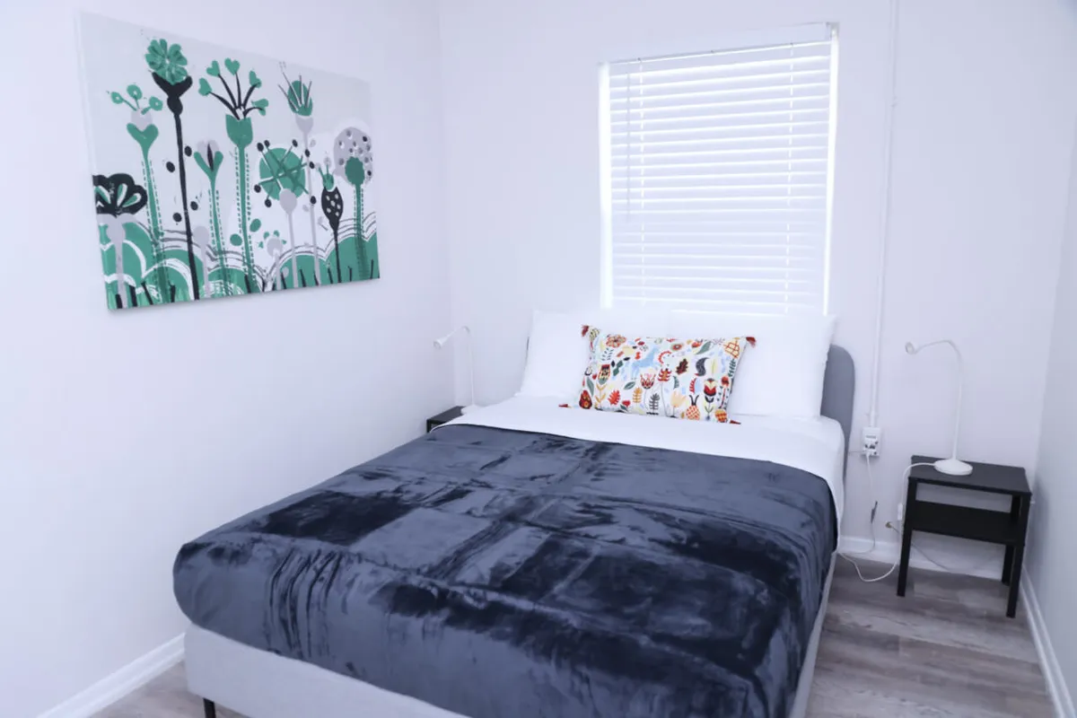 A peaceful sleeping area with a bed dressed in a dark blue blanket and colorful pillow, a window with white blinds allowing natural light, green-themed abstract wall art, and a black nightstand with a reading lamp. The scene exudes calm and restfulness—ideal for relaxation after a busy day.