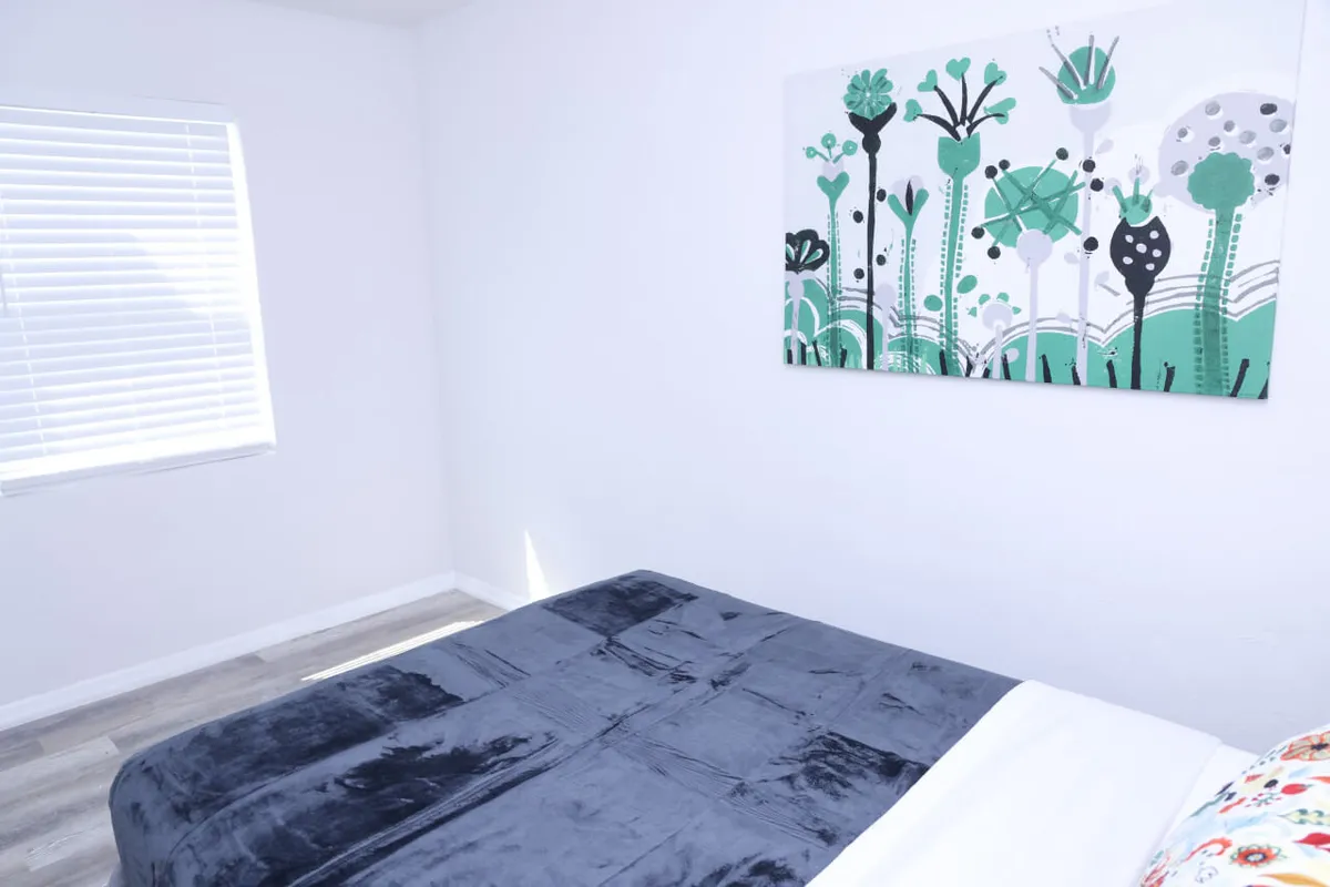 A cozy, sunlit bedroom with a plush dark gray bedspread, abstract botanical wall art, and bright white walls. Natural light streams through a window, creating a fresh and inviting atmosphere.