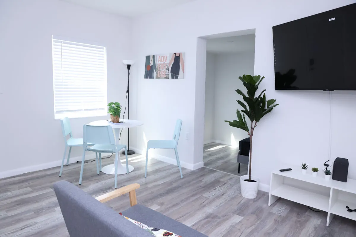 A bright, inviting studio space featuring a round white dining table set with light blue chairs, a large flat-screen TV mounted on the wall, a comfortable gray sofa with a visible wooden armrest, and a potted green plant near the doorway. Natural light fills the room, enhancing the modern minimalist décor and creating a fresh, homey atmosphere.