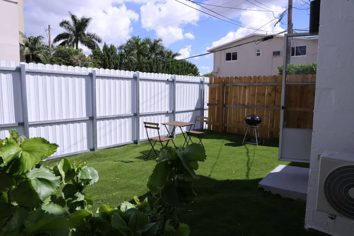 A small, private patio with a wooden table and two chairs, a charcoal BBQ grill, and a green grass lawn, all enclosed by a white fence. The space feels relaxed and inviting, perfect for outdoor dining or a quiet coffee.